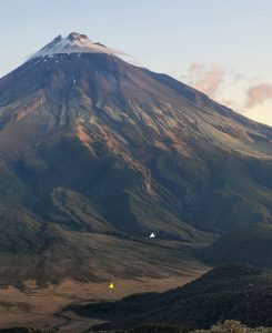 One of the many mountain streams that drain the slopes of Mt Taranaki merging into a small, lower gradient alluvial fan near the base of the edifice. The outer edge of the fan (yellow arrow) interfingers with a swamp (brown hues). Unlike the mountain torrents, the alluvial fan is characterised by gravel bars and bedforms, and an improvement in clast sorting. Holly Hut indicated by the white arrow. Pouakai track, Mt. Taranaki, New Zealand. Photo by Sam Ricketts. 