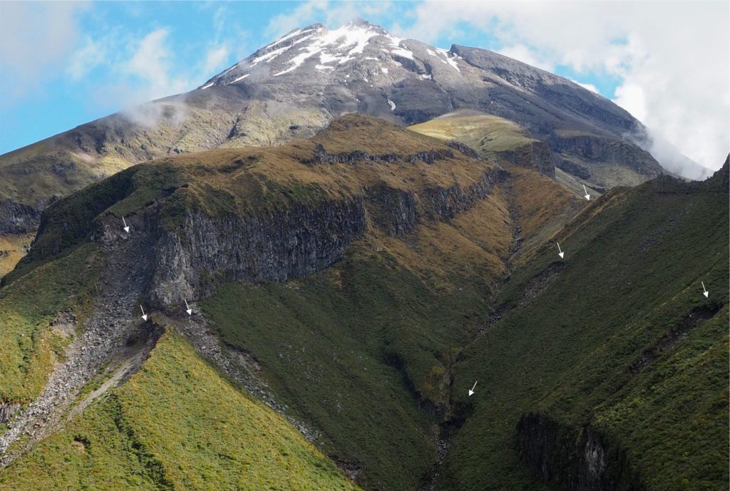 Bluffs that expose stacked, columnar jointed lava flows are a prime source of blocky fragments that tumble or slide directly into the narrow channel or become part of the slope colluvium. Supply of debris to the channel also occurs via gravitational failure of the colluvium – seven recent failures and their head scarps are visible here (arrows). The main bluff (left-centre) is about 75 m high. Holly Hut track, Mt. Taranaki, New Zealand.